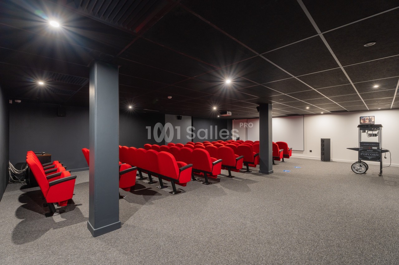 Salle de projection avec rangées de sièges rouges, écran blanc à l'arrière et équipement audiovisuel sur la droite.