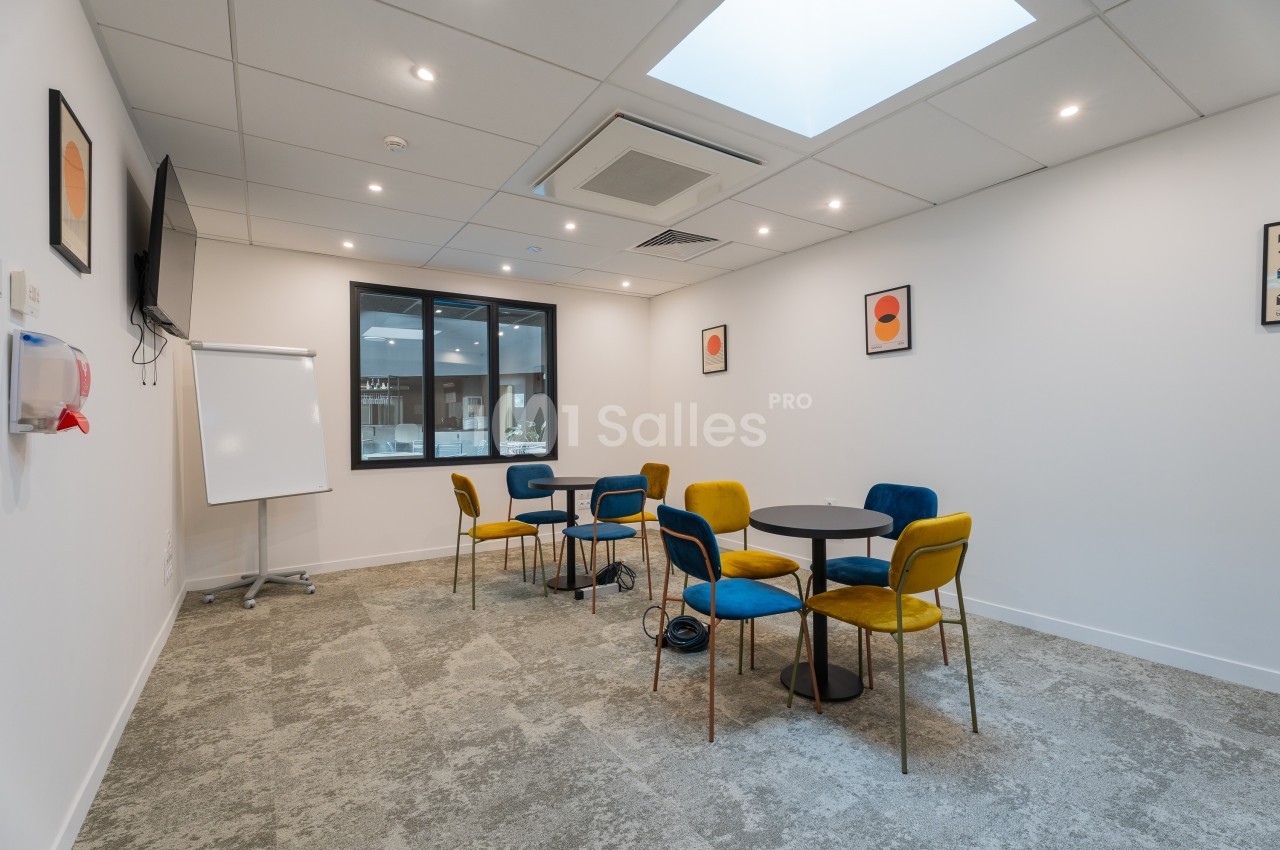 Salle de réunion lumineuse avec tables rondes, chaises colorées, tableau blanc et fenêtre donnant sur un autre espace.