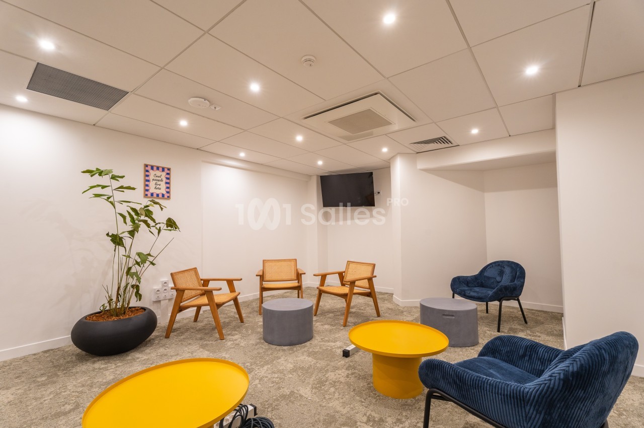 Salle lumineuse avec fauteuils en bois et en velours, tables basses jaunes et grises, et une plante en pot.