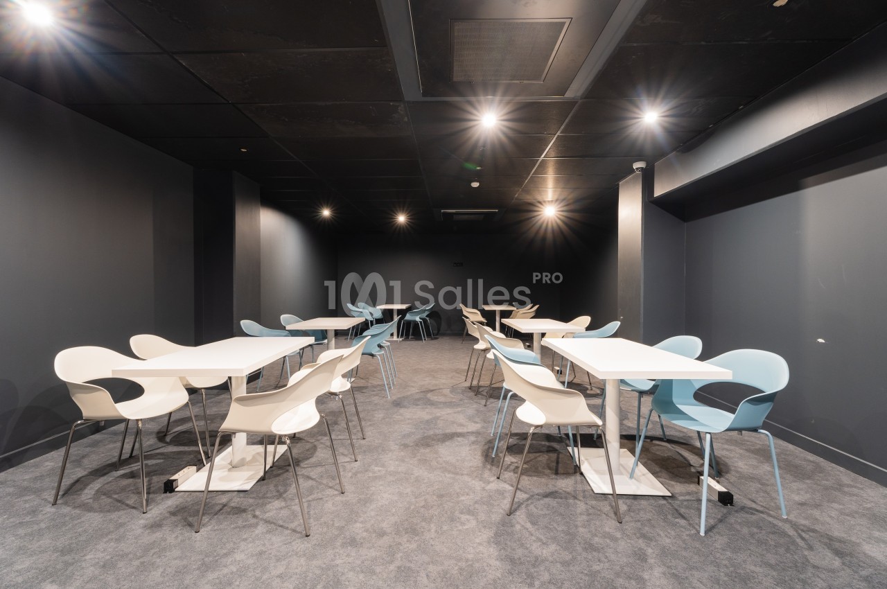 Salle moderne avec des murs sombres, éclairage au plafond, tables blanches et chaises design beige et bleues.