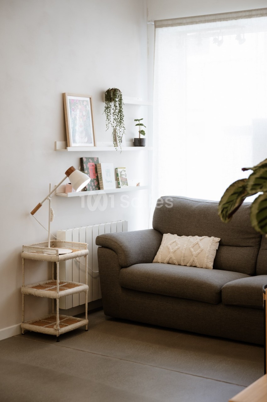 Salon lumineux avec canapé gris, étagères murales décorées, lampe sur table et grande fenêtre avec rideau clair.