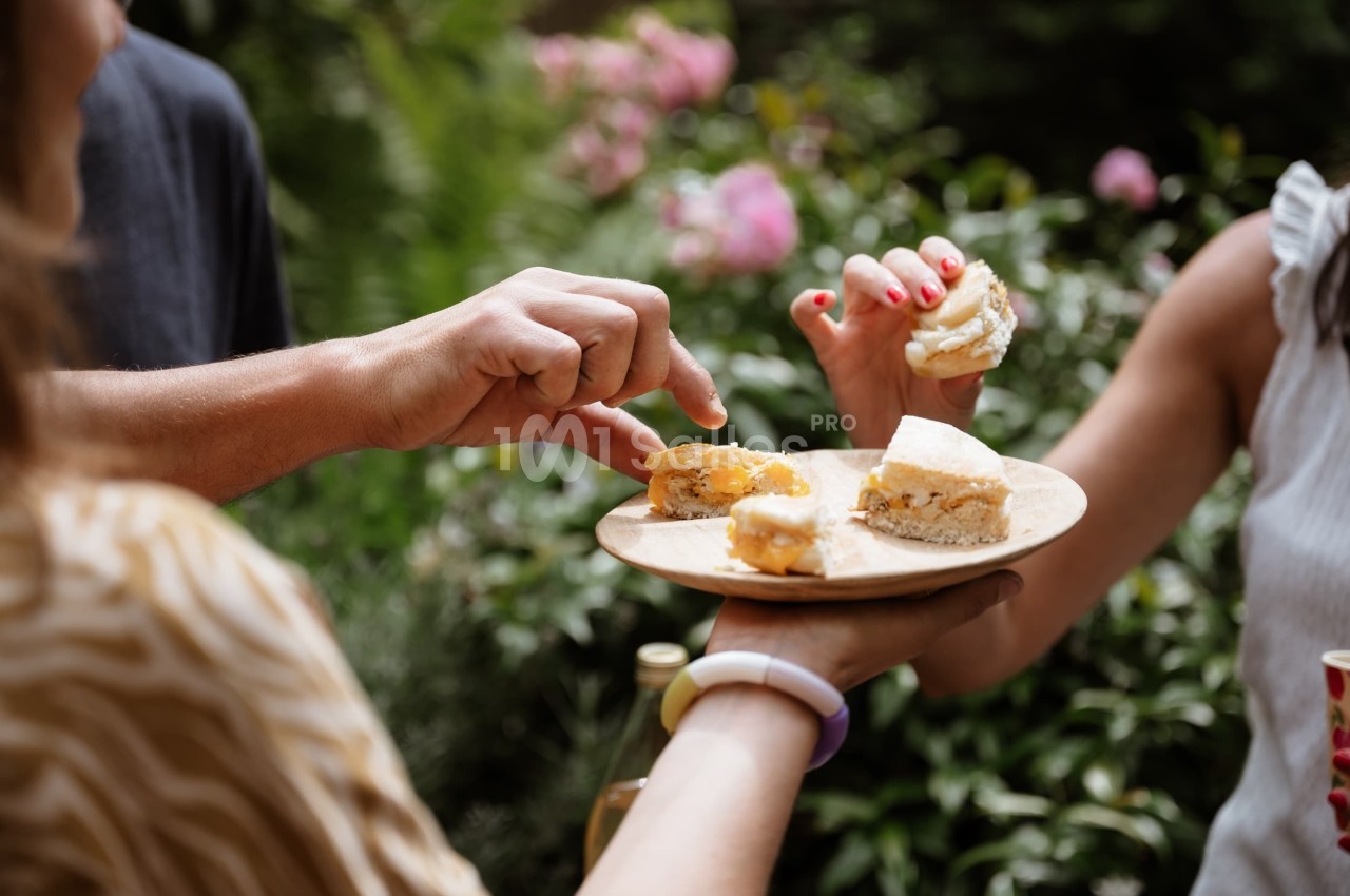 Des mains partagent des amuse-bouches sur une assiette en bois dans un jardin verdoyant.