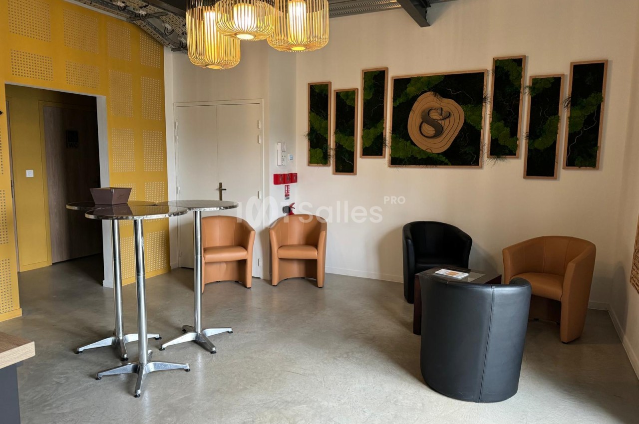 Espace de réception aéré avec tables hautes, fauteuils caramel et noirs, luminaires dorés, murs blancs et panneaux jaunes.