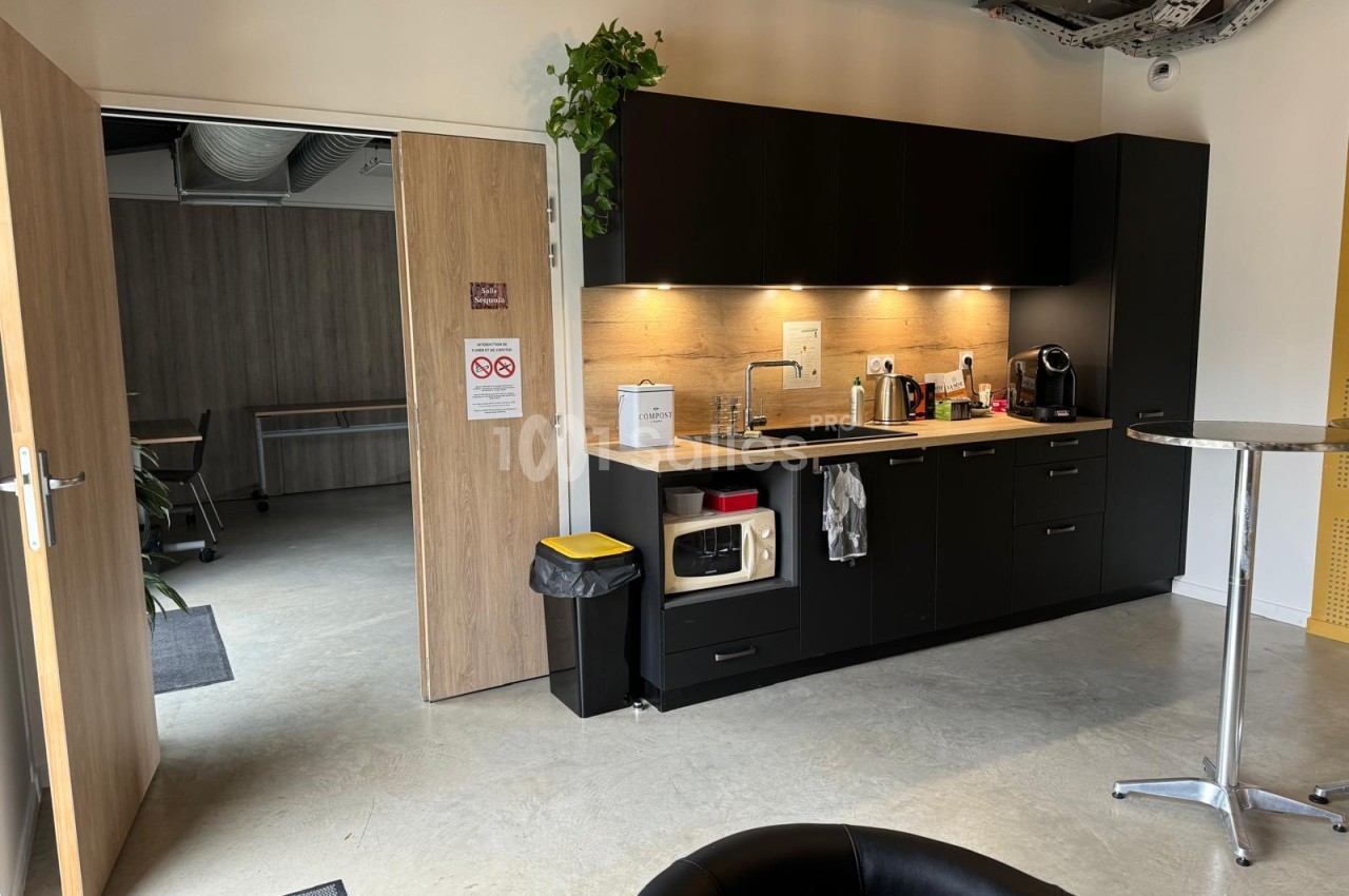 Cuisine équipée avec mobilier noir et plan de travail bois, accès à salle de réunion visible à gauche.