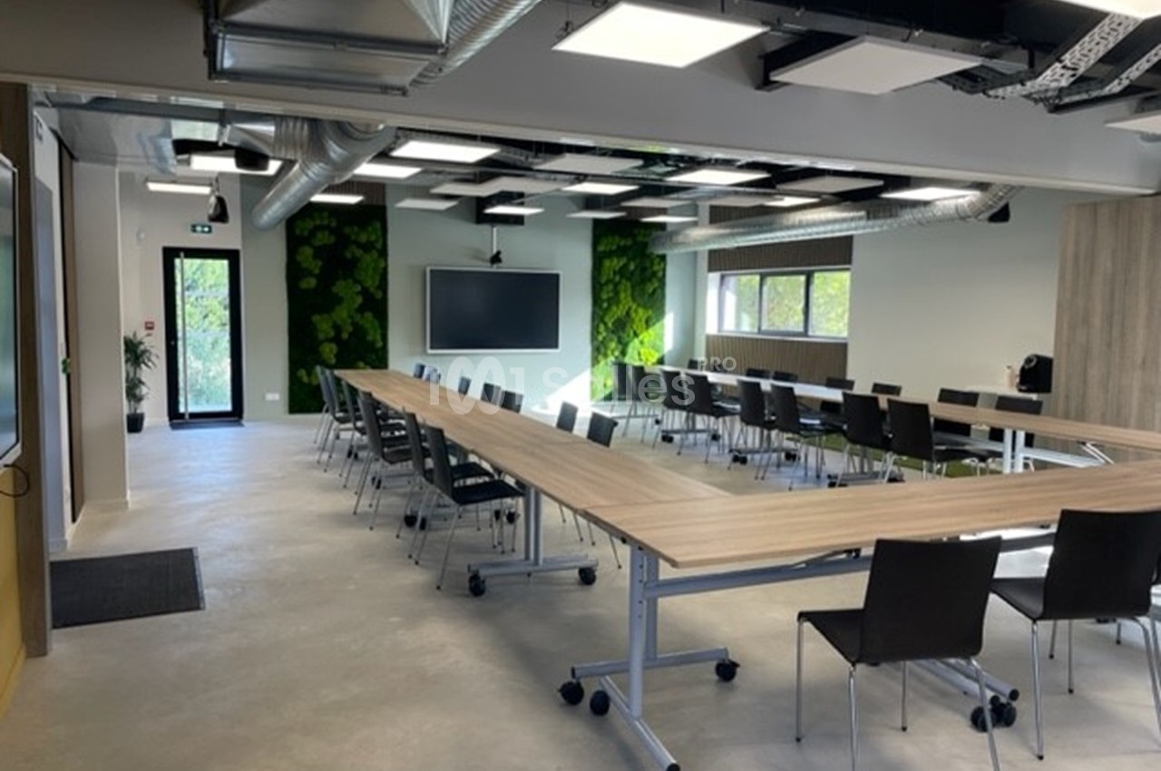 Salle de réunion moderne avec tables en bois, chaises noires sur roulettes, écran mural et murs végétalisés verts.