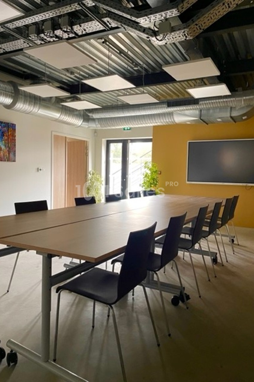 Salle de réunion équipée d'une grande table rectangulaire, chaises noires mobiles, écran, mur ocre et plafond avec gaines d'aération.