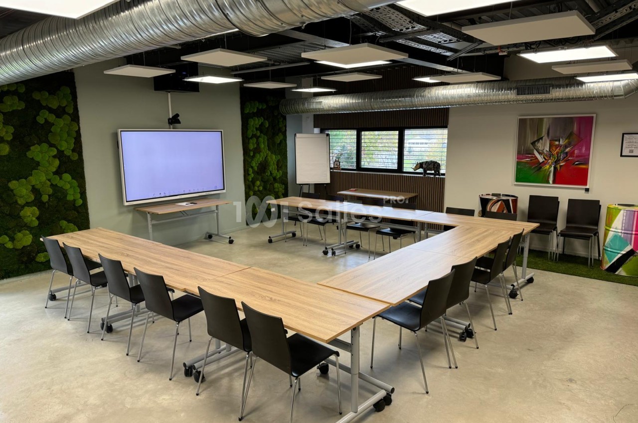 Salle de réunion avec table en U en bois clair, chaises noires, écran de présentation, tuyauterie apparente et murs décorés de plantes vertes.