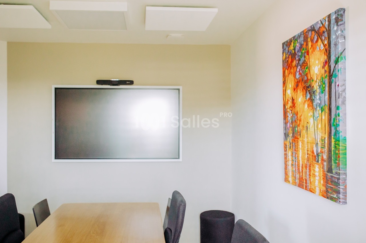 Salle de réunion avec écran plat mural, tableau coloré et table en bois avec chaises grises.