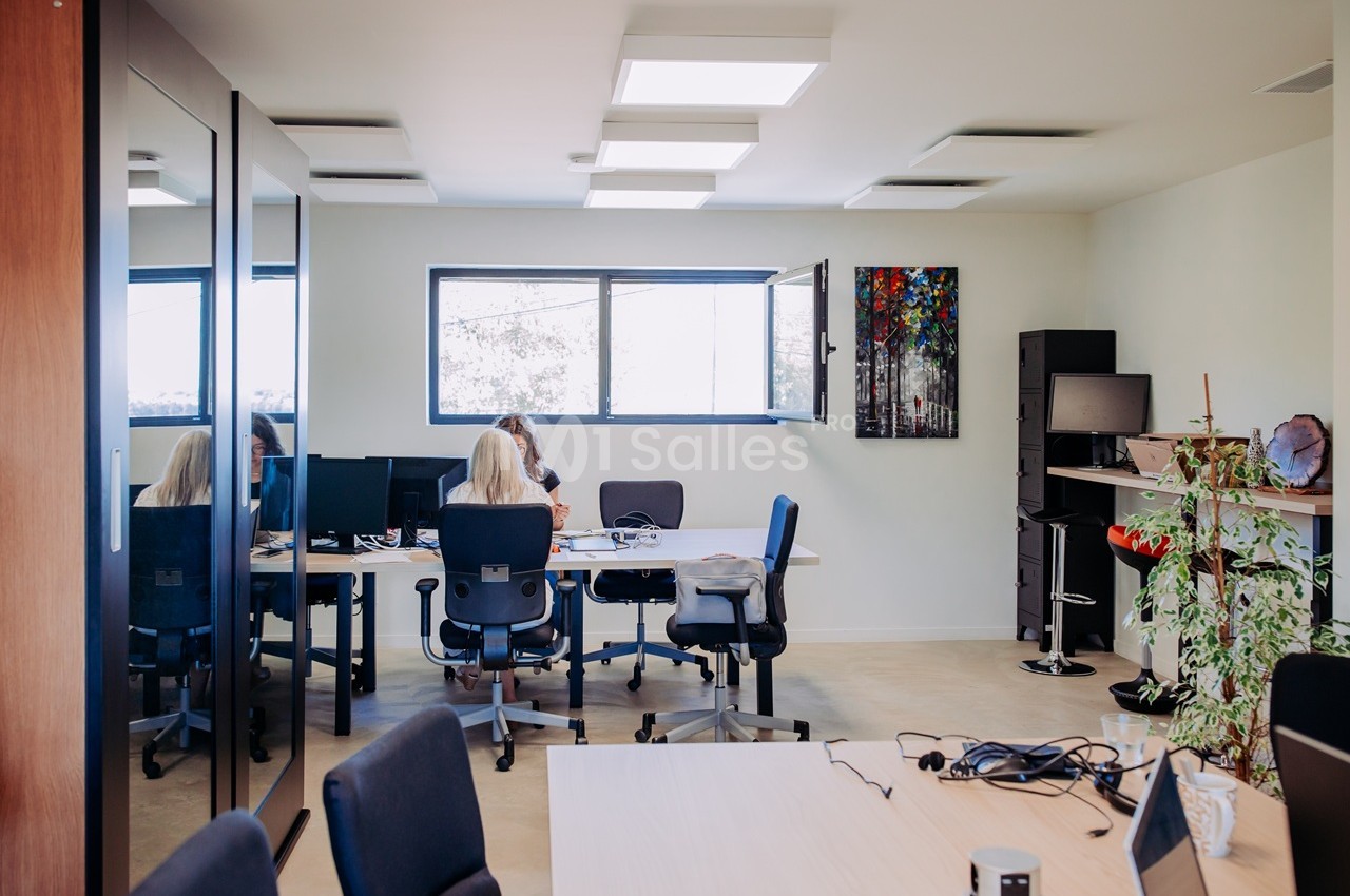 Espace de coworking ouvert avec bureaux équipés, chaises noires, grandes fenêtres et éclairage zénithal.