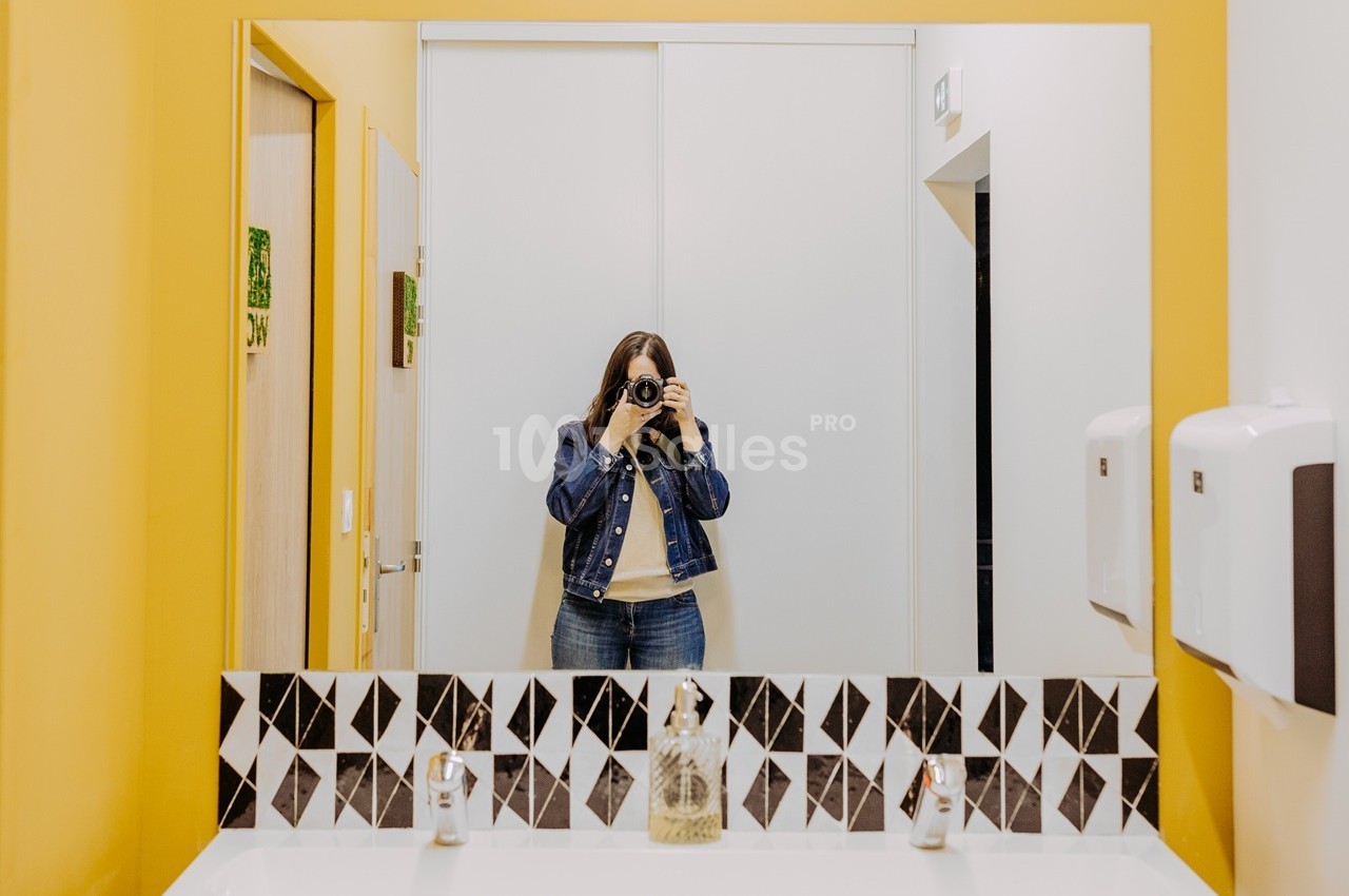 Salle avec murs jaunes et blancs, miroir encadré de carreaux géométriques noir et blanc, objets de décoration sur le rebord.