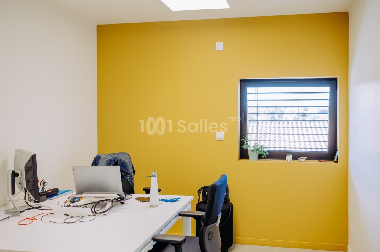 Espace de travail avec mur jaune vif, bureau blanc, chaise de bureau bleue et noire, équipement informatique et fenêtre avec store.