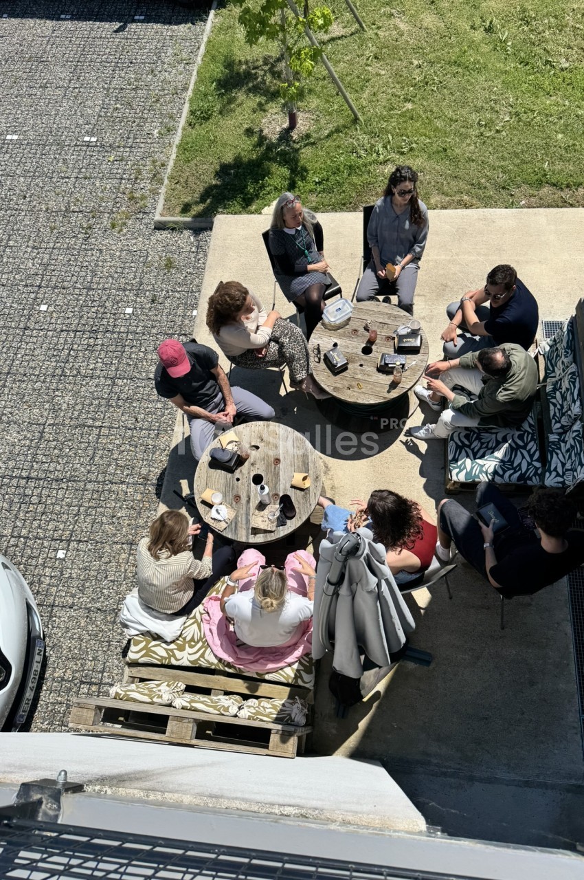 Terrasse extérieure avec deux tables rondes en bois accueillant environ dix personnes assises, entourée de pavés et de gazon.
