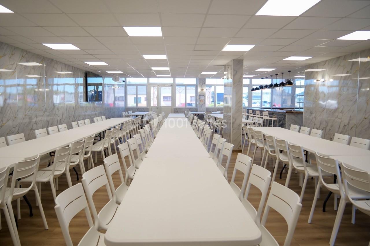 Salle lumineuse avec de longues tables blanches alignées et des chaises en plastique, entourées de murs clairs.