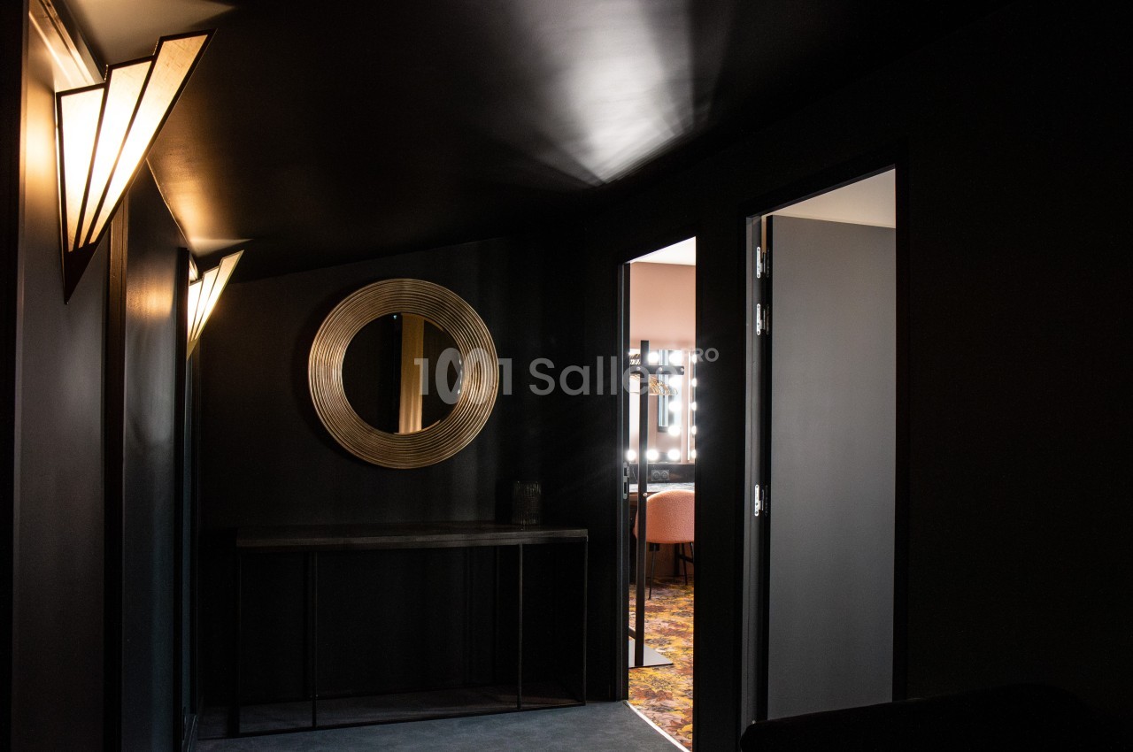 Couloir sombre avec un miroir rond doré, une console noire et des portes ouvertes laissant entrevoir des pièces éclairées.