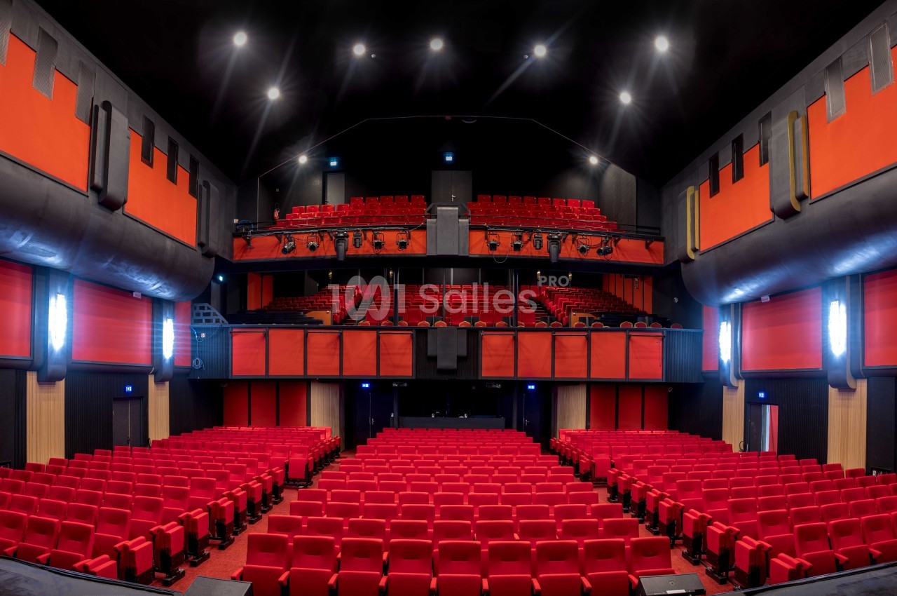 Vue intérieure d'une salle de théâtre moderne avec des rangées de sièges rouges et une scène au fond.