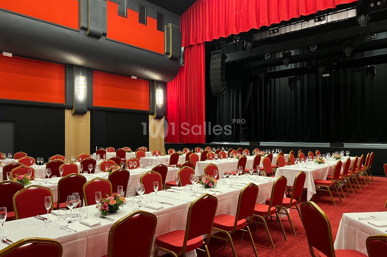 Salle de réception avec tables dressées, nappes blanches, chaises rouges et scène en arrière-plan avec rideaux rouges.