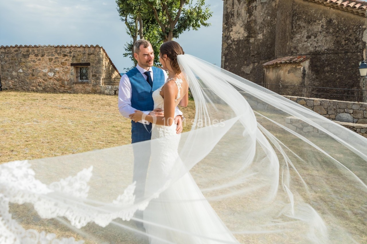 Un couple en tenue de mariage se tient dans un jardin près d'un bâtiment en pierre, avec un voile flottant au vent.