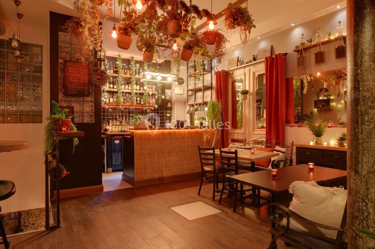 Intérieur d'un restaurant chaleureux avec des plantes suspendues, un éclairage tamisé et un bar en arrière-plan.
