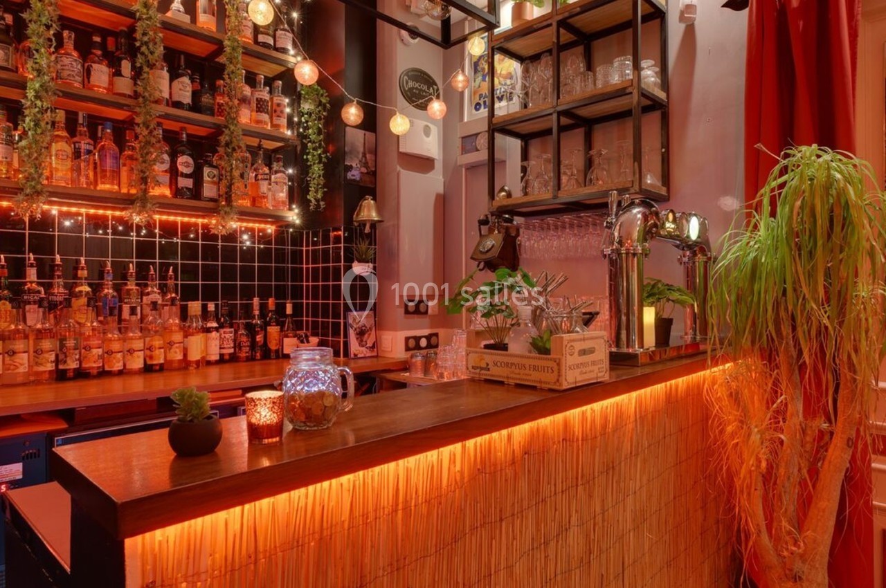 Bar chaleureux avec comptoir en bois, étagères garnies de bouteilles, lumières tamisées et décorations végétales.