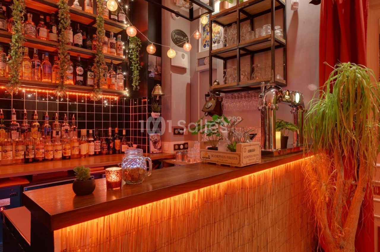 Bar chaleureux avec comptoir en bois, étagères remplies de bouteilles, lumières tamisées et décorations végétales.