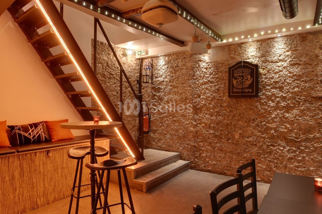Espace intérieur chaleureux avec murs en pierre, escalier éclairé, table haute et banquette avec coussins colorés.