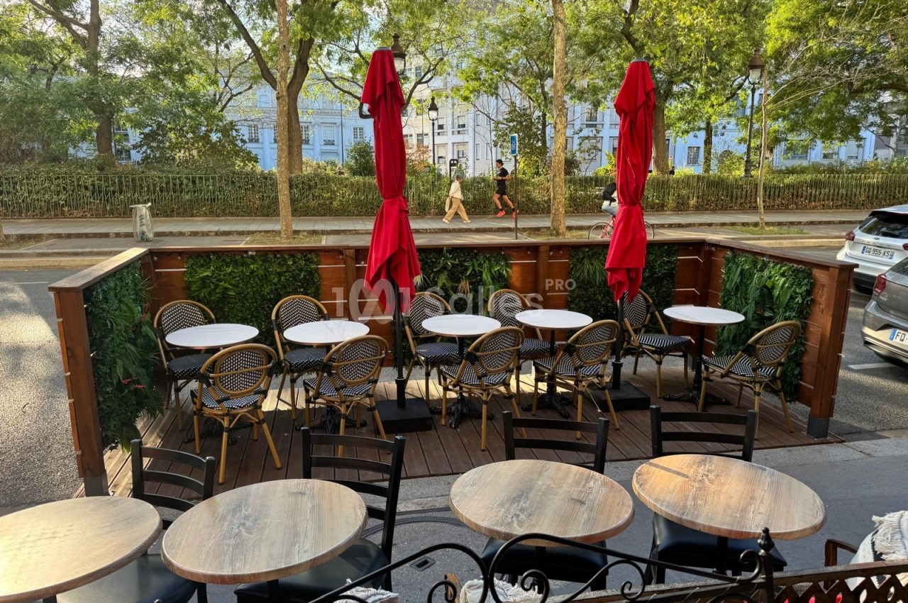 Terrasse extérieure avec tables rondes, chaises en rotin et parasols rouges, bordée de verdure et donnant sur une rue…