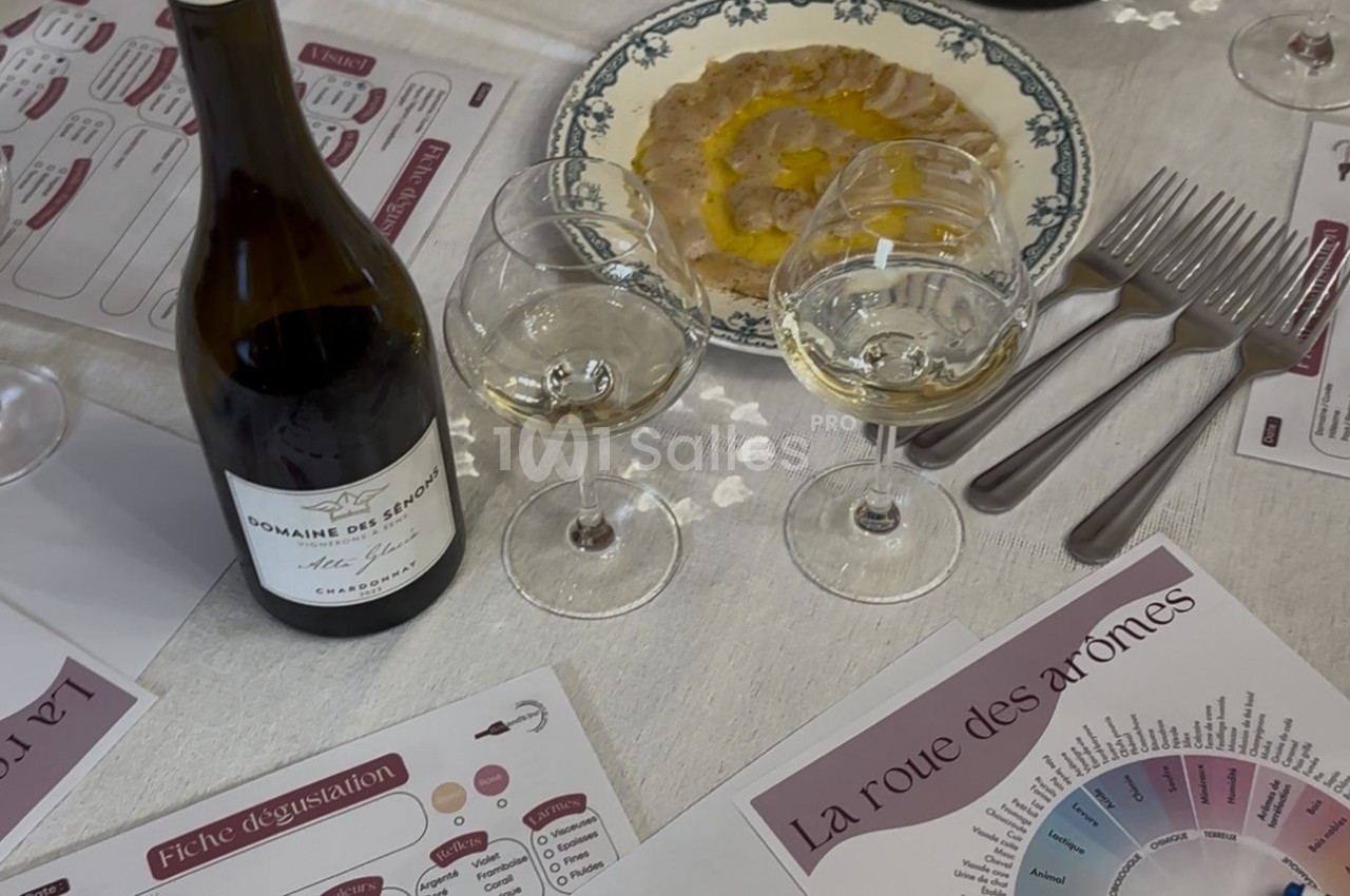 Bouteille de vin, verres remplis, assiette de carpaccio et documents de dégustation sur une table dressée.