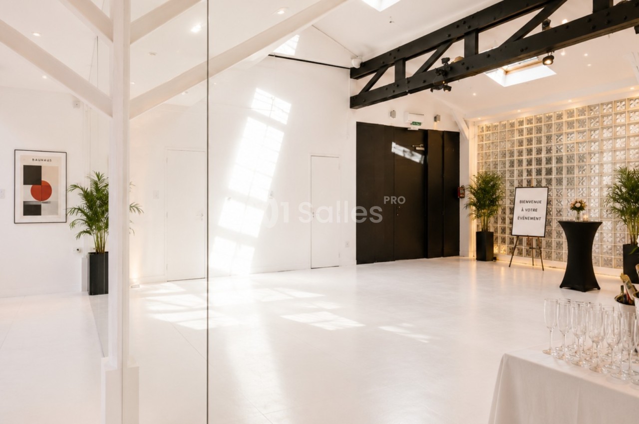 Salle lumineuse avec murs blancs, verrière, poutres noires apparentes, plantes décoratives et table avec flûtes de champagne.