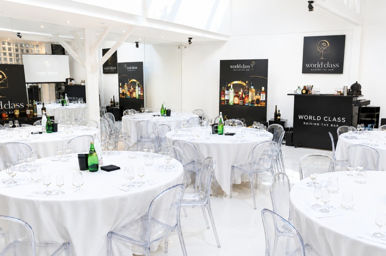 Salle lumineuse avec des tables rondes dressées, des chaises transparentes et des affiches ’World Class’ en arrière-plan.