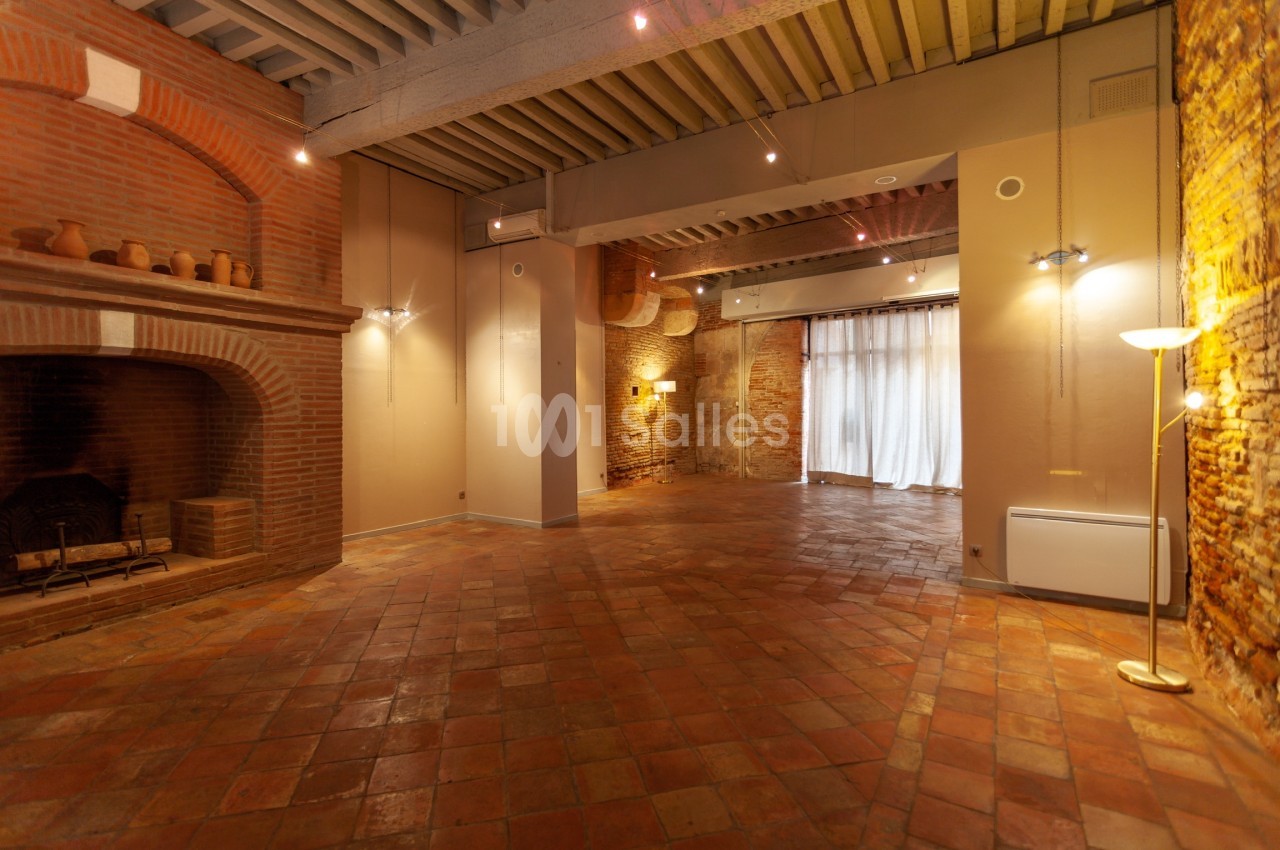 Salle vide avec sol en terre cuite, murs en briques, grande cheminée et lumière naturelle entrant par des rideaux.