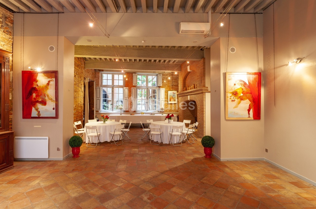 Salle lumineuse avec murs en briques, tables rondes dressées, chaises blanches et grandes fenêtres donnant sur un jardin.