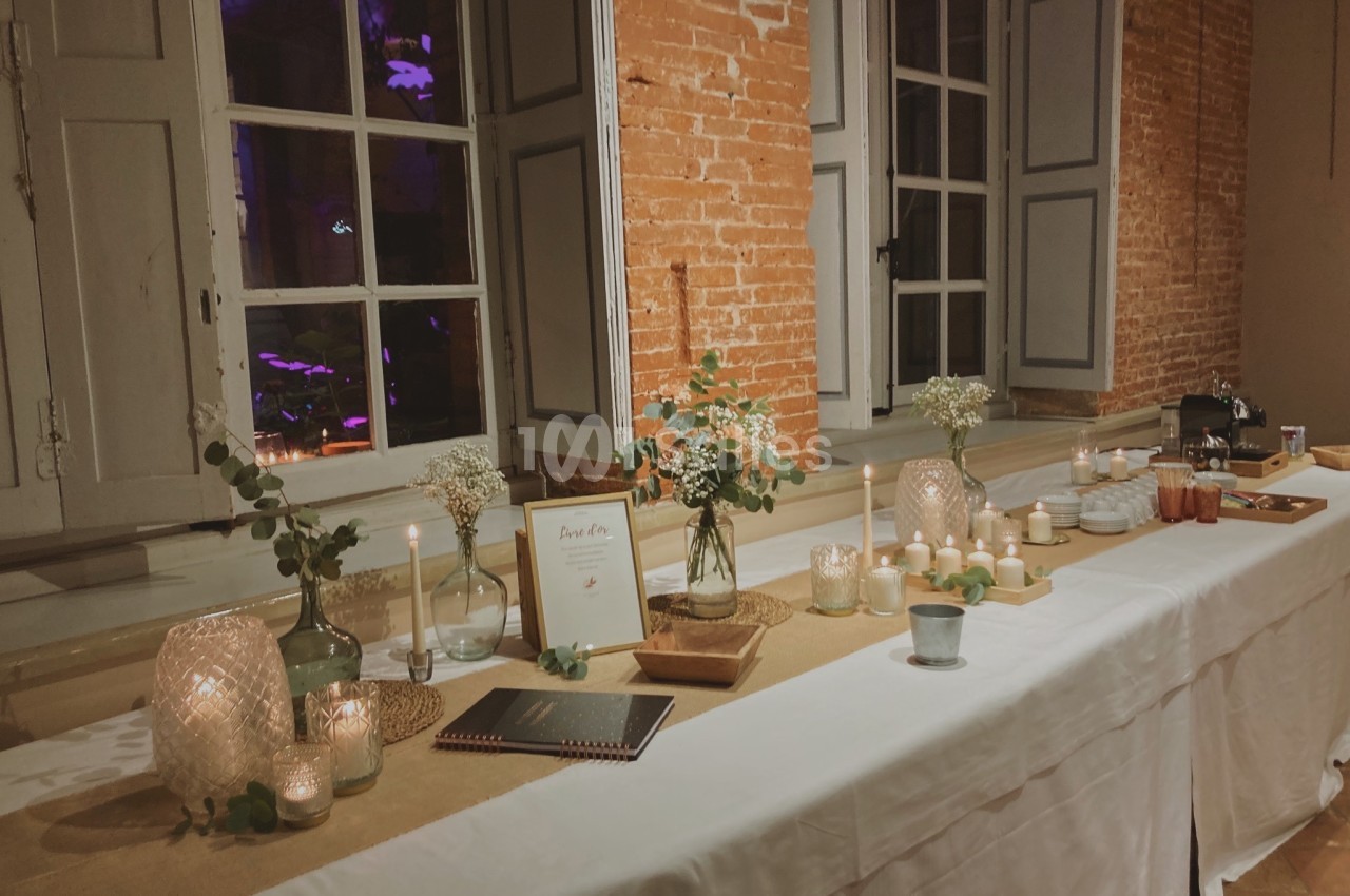 Table décorée avec bougies, fleurs et cadres, placée devant un mur en briques et des fenêtres ouvertes.