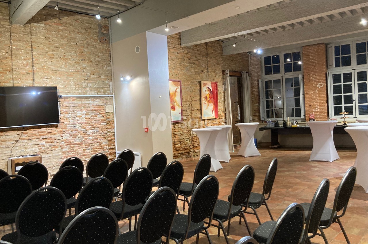 Salle avec murs en briques, rangées de chaises noires, tables hautes blanches et écran mural, éclairée par des spots.