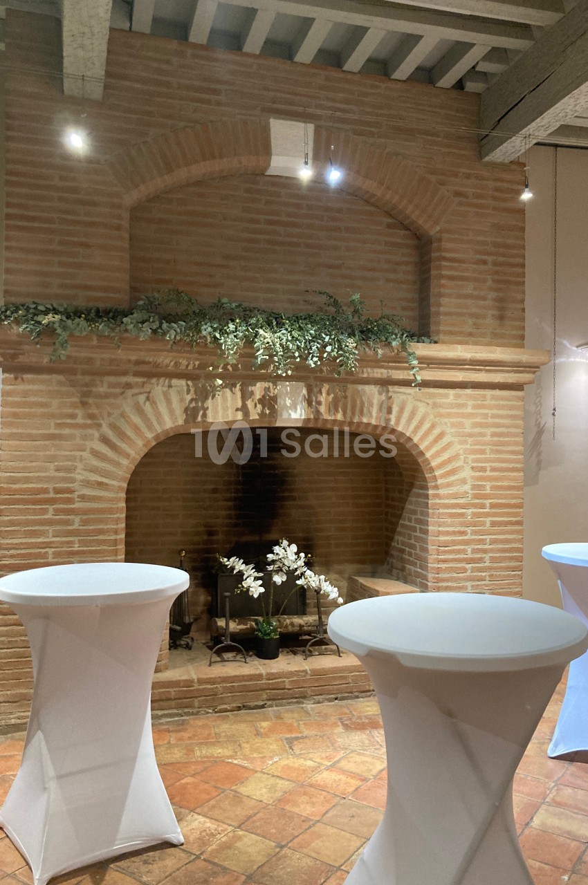 Cheminée en briques décorée de plantes, entourée de tables hautes drapées de blanc dans une salle au sol en terre cuite.