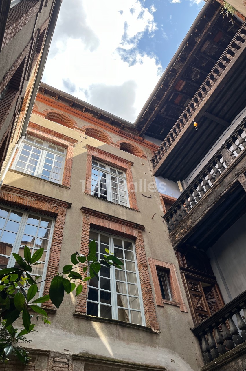 Cour intérieure avec façades en briques, grandes fenêtres et balcons en bois sous un ciel partiellement nuageux.