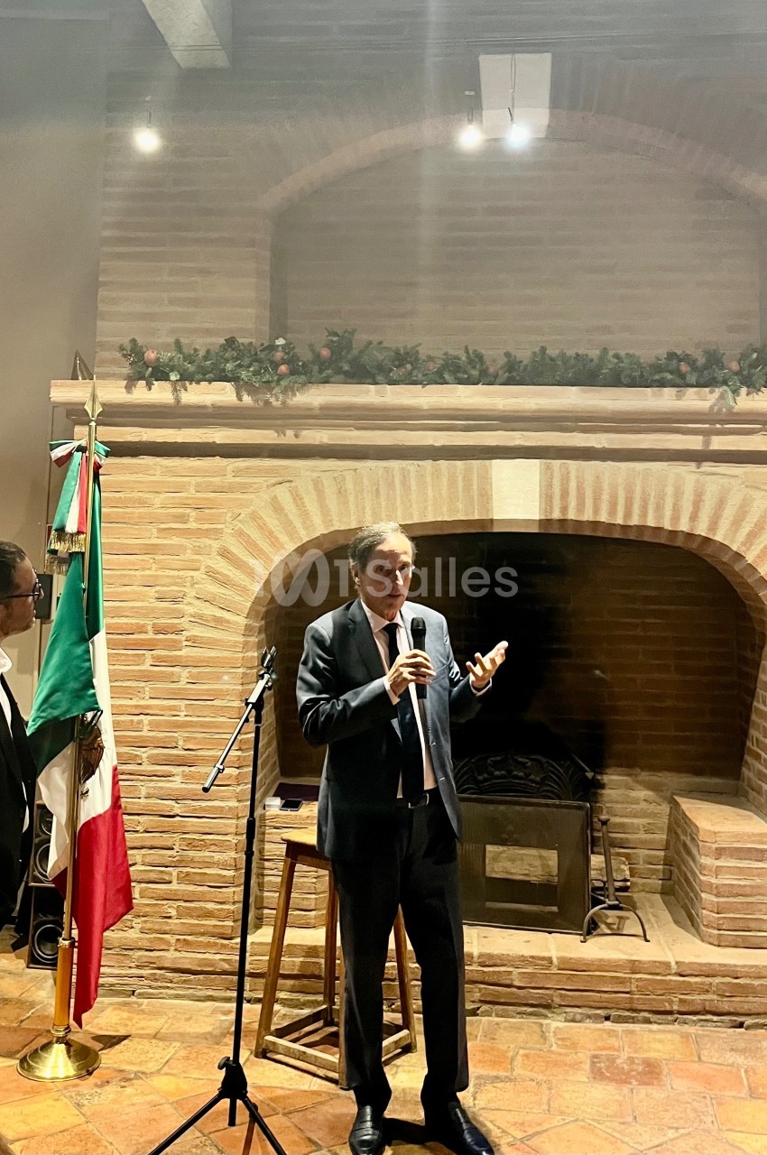 Un homme en costume parle au micro devant une cheminée en briques, près des drapeaux français et mexicain.
