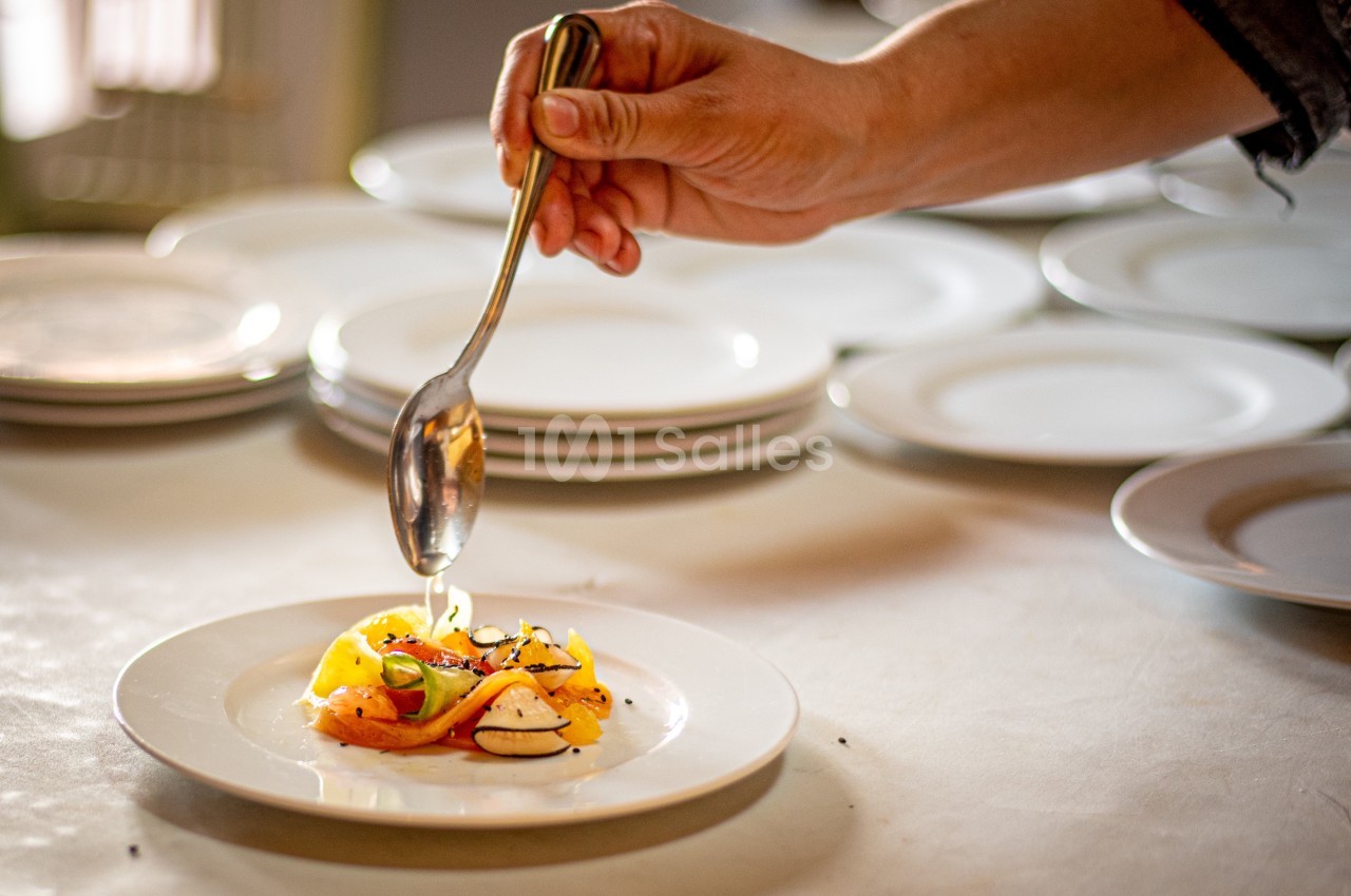 Une main dresse une assiette avec des légumes colorés et une cuillère, dans une cuisine avec des assiettes en arrière-plan.