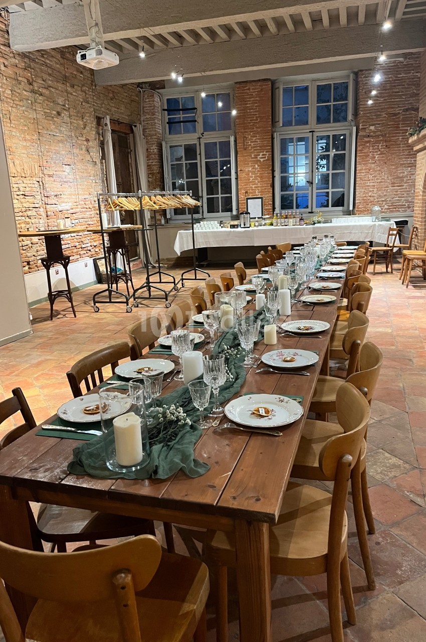 Grande table en bois dressée avec vaisselle, bougies et décorations, dans une salle au mur de briques et sol en terre cuite.