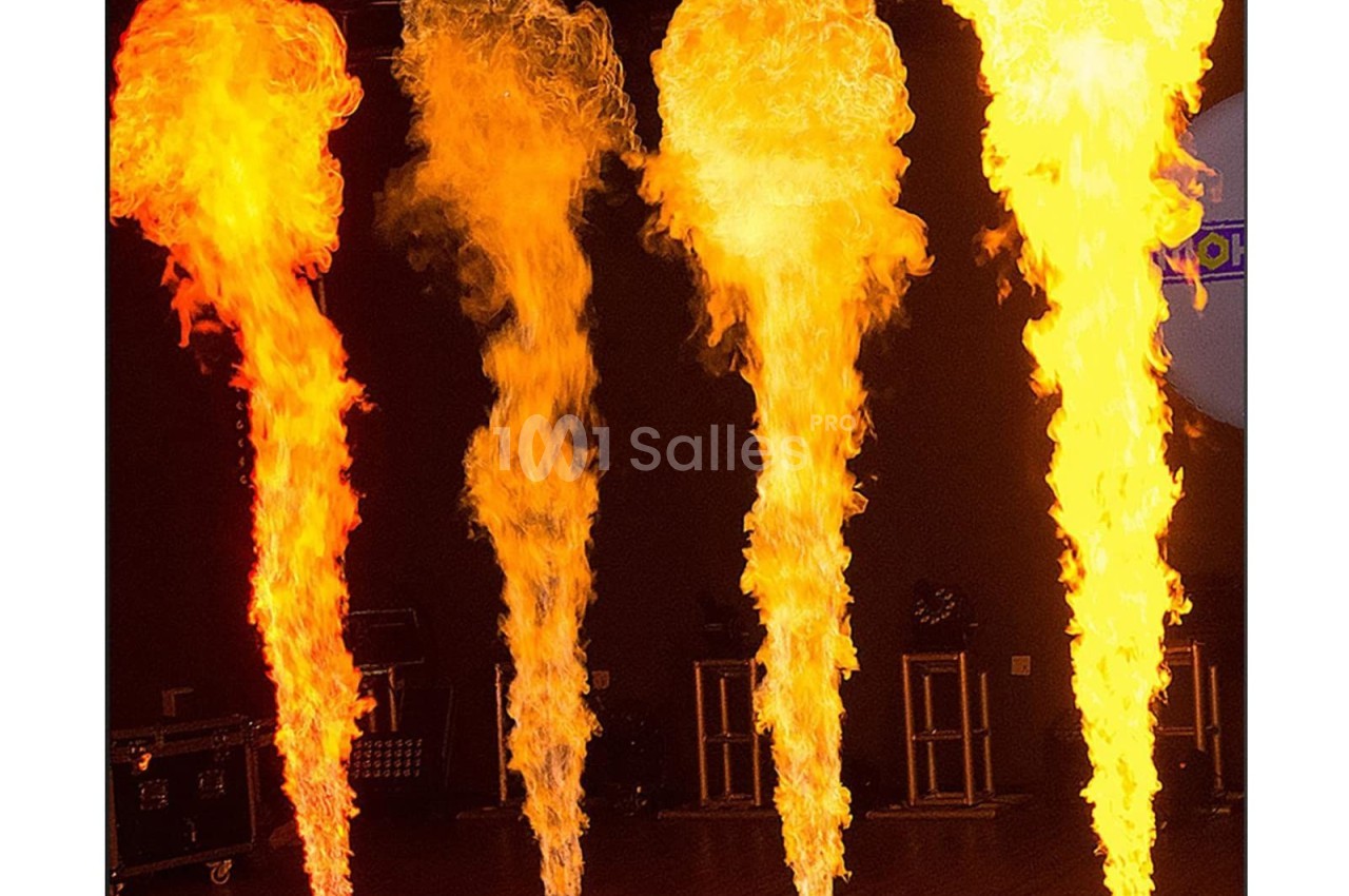 Quatre colonnes de flammes jaillissent de dispositifs au sol dans une salle sombre.