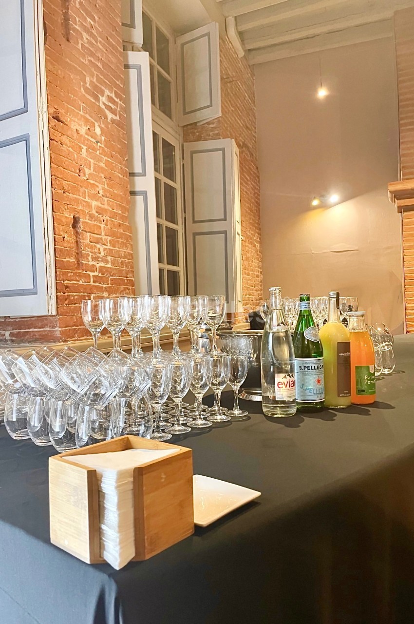 Table avec verres empilés, bouteilles d'eau et de jus, dans une pièce aux murs en briques et grandes fenêtres.