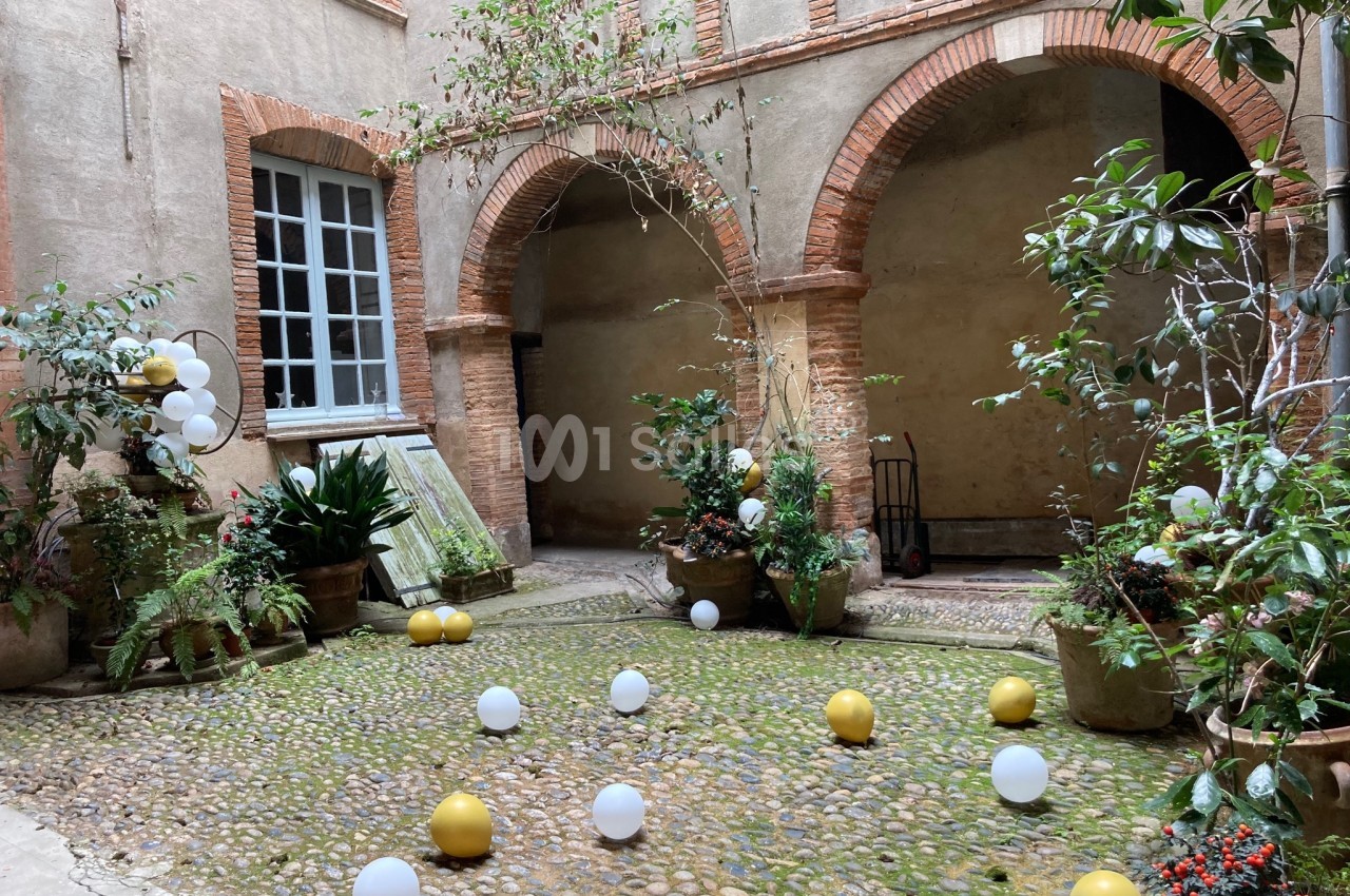 Cour intérieure pavée avec des arches en briques, des plantes en pots et des ballons blancs et dorés dispersés au sol.