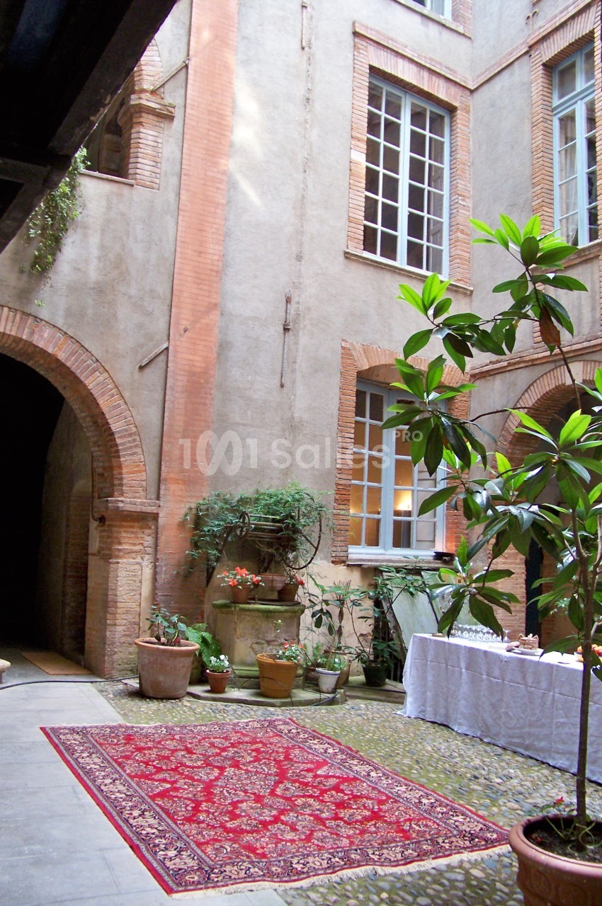 Cour intérieure avec murs en briques, tapis rouge, plantes en pot et table dressée sous une lumière naturelle.