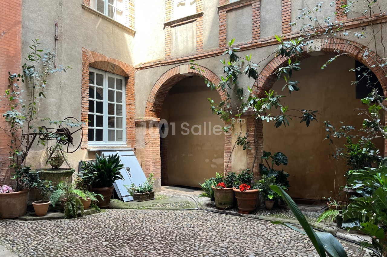 Cour intérieure avec pavés, plantes en pots, arches en briques et mur beige, éclairée par une lumière naturelle.