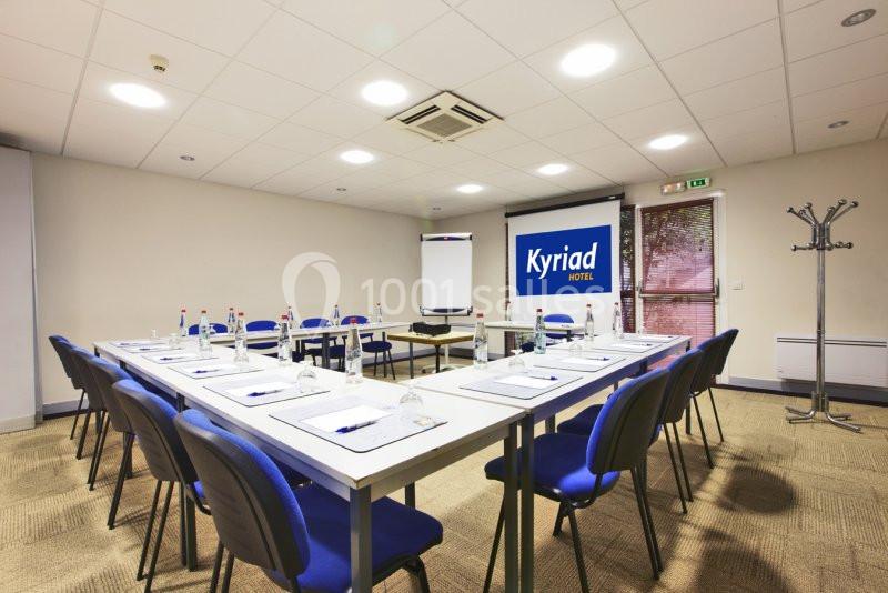 Salle de réunion équipée avec tables, chaises, paperboard, bouteilles d'eau et écran affichant ’Kyriad Hotel’.