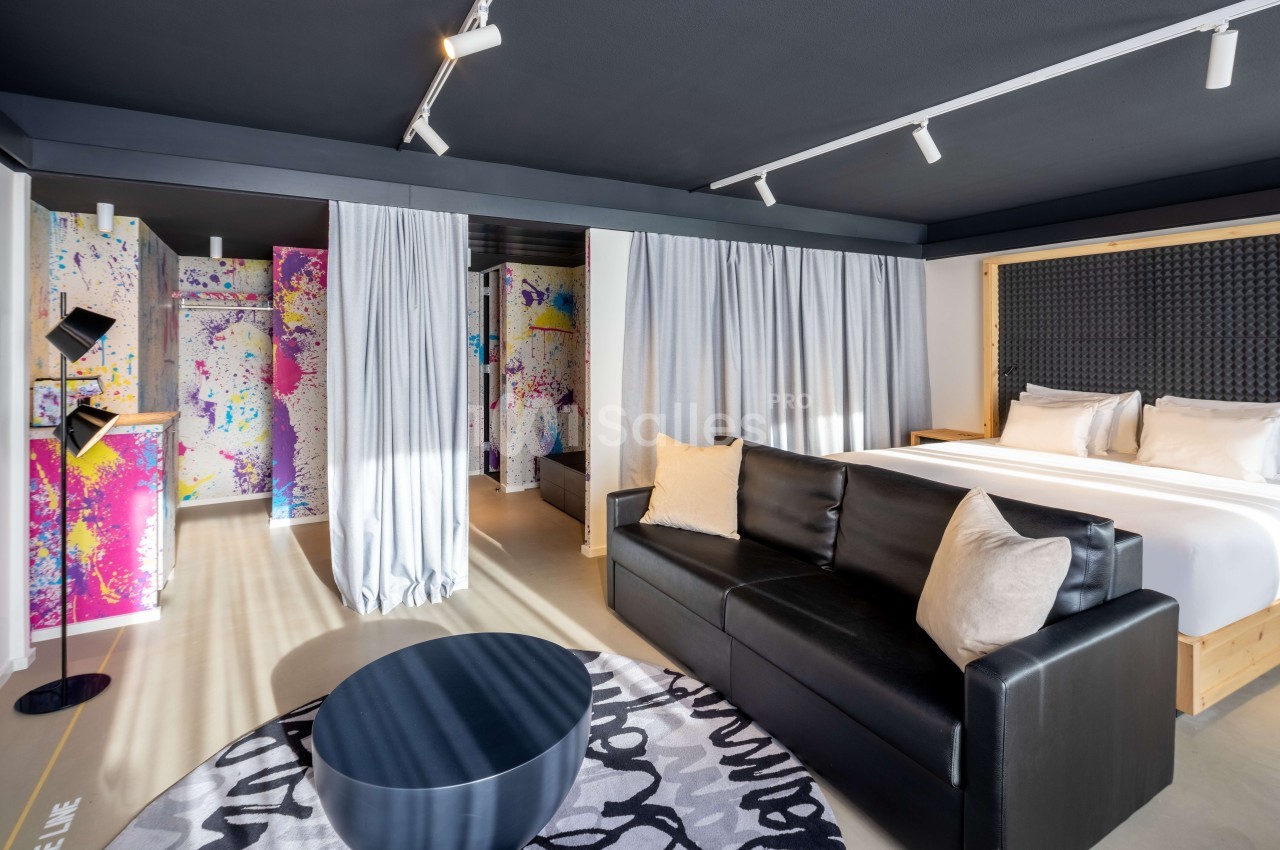Suite hôtel moderne avec lit queen-size, canapé noir, murs aux motifs colorés et rideaux blancs coulissants.
