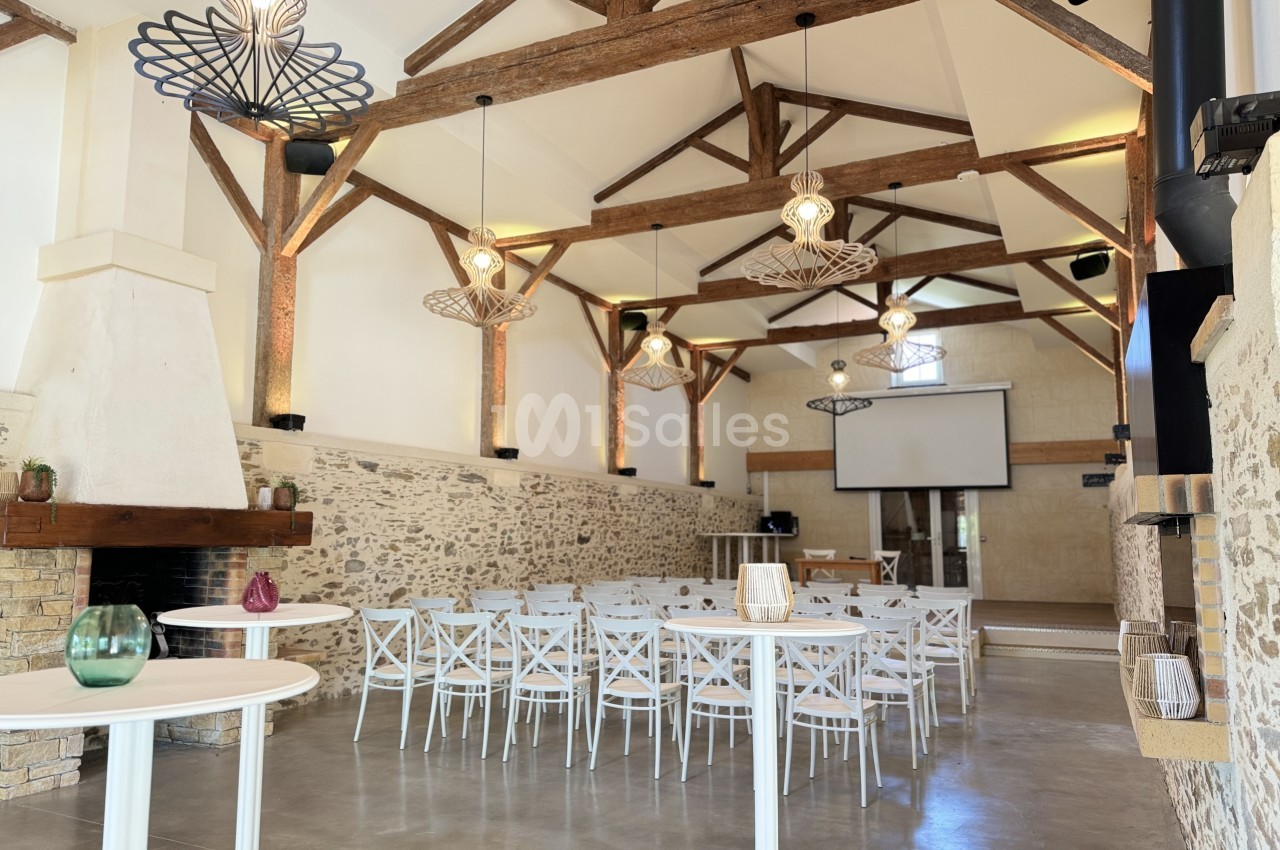 Salle avec charpente bois apparente, lustre design géométrique, chaises blanches, murs pierre et foyer - intérieur spacieux et lumineux.