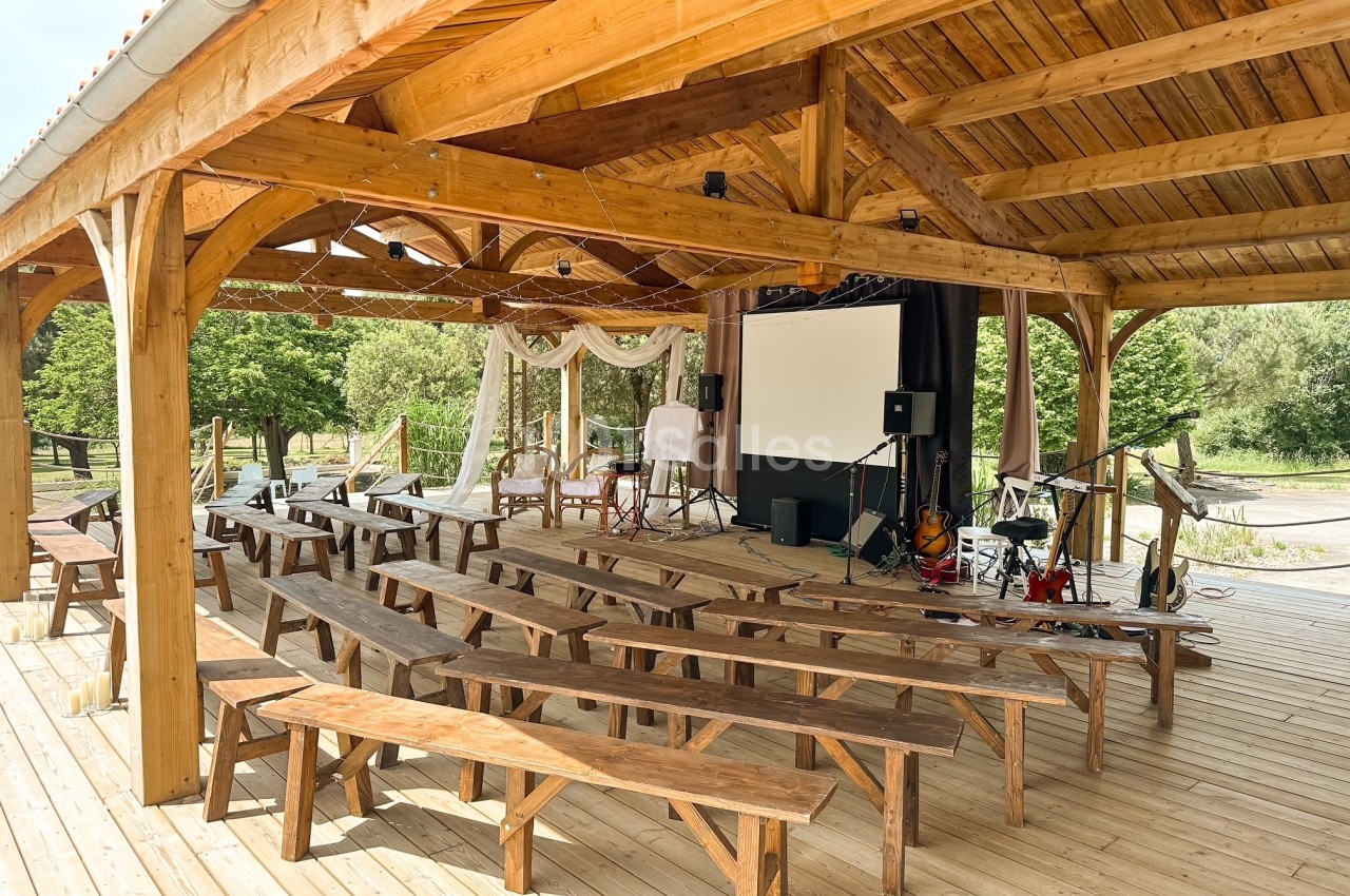 Charpente en bois abritant des tables et bancs en bois face à une scène équipée d'un écran de projection et instruments de musique.