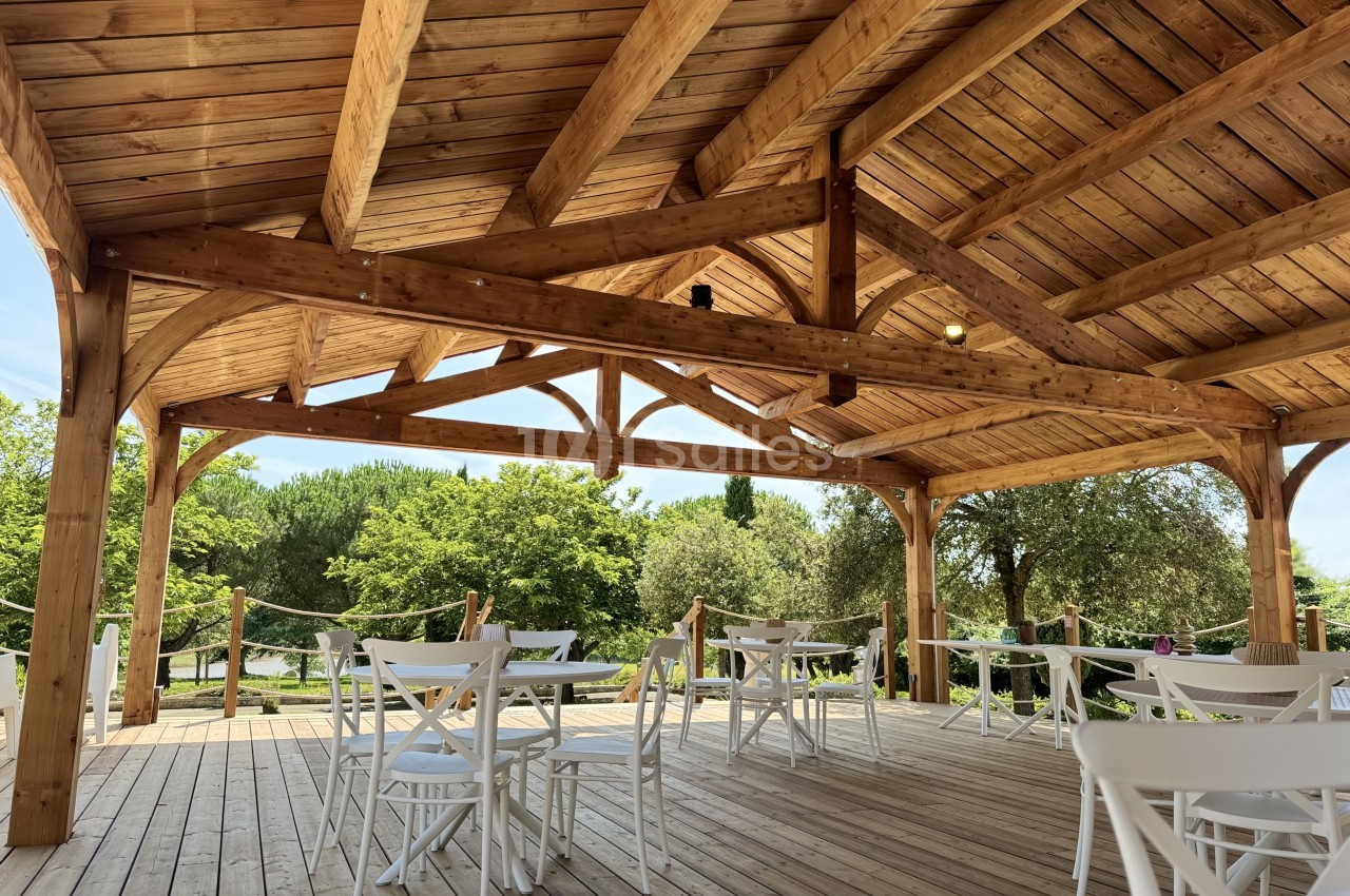 Terrasse couverte en bois avec charpente apparente, chaises blanches et tables, entourée de verdure.
