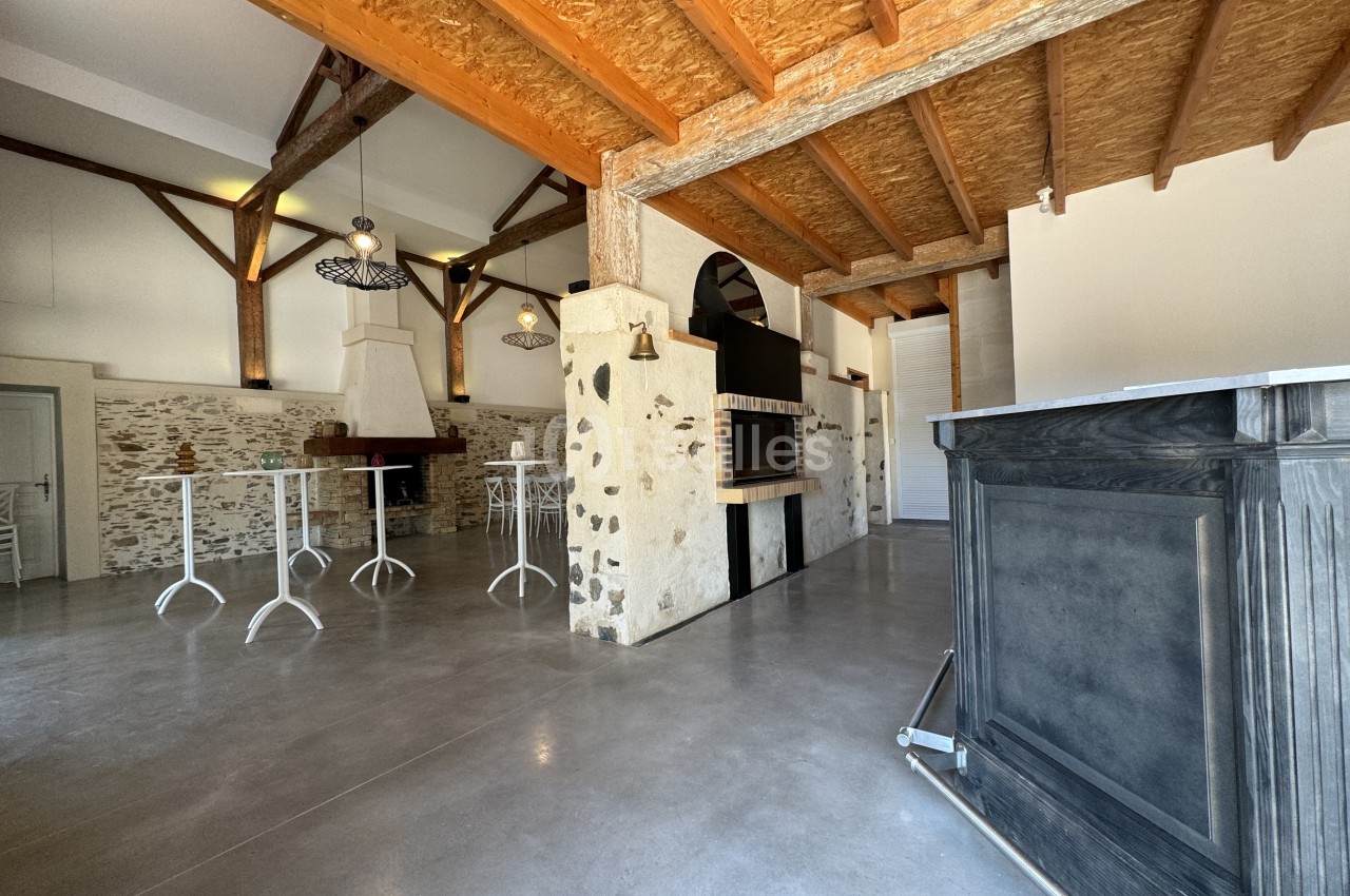 Salle vide avec poutres apparentes, comptoir en pierre, tables hautes et bar gris, sol béton brut.