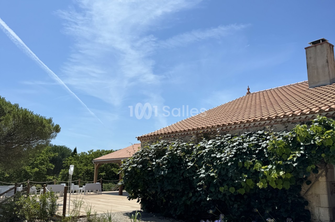 Bâtisse en pierre avec toiture en tuiles, lierre blanc grimpant sur la façade, terrasse dallée et mobilier blanc.